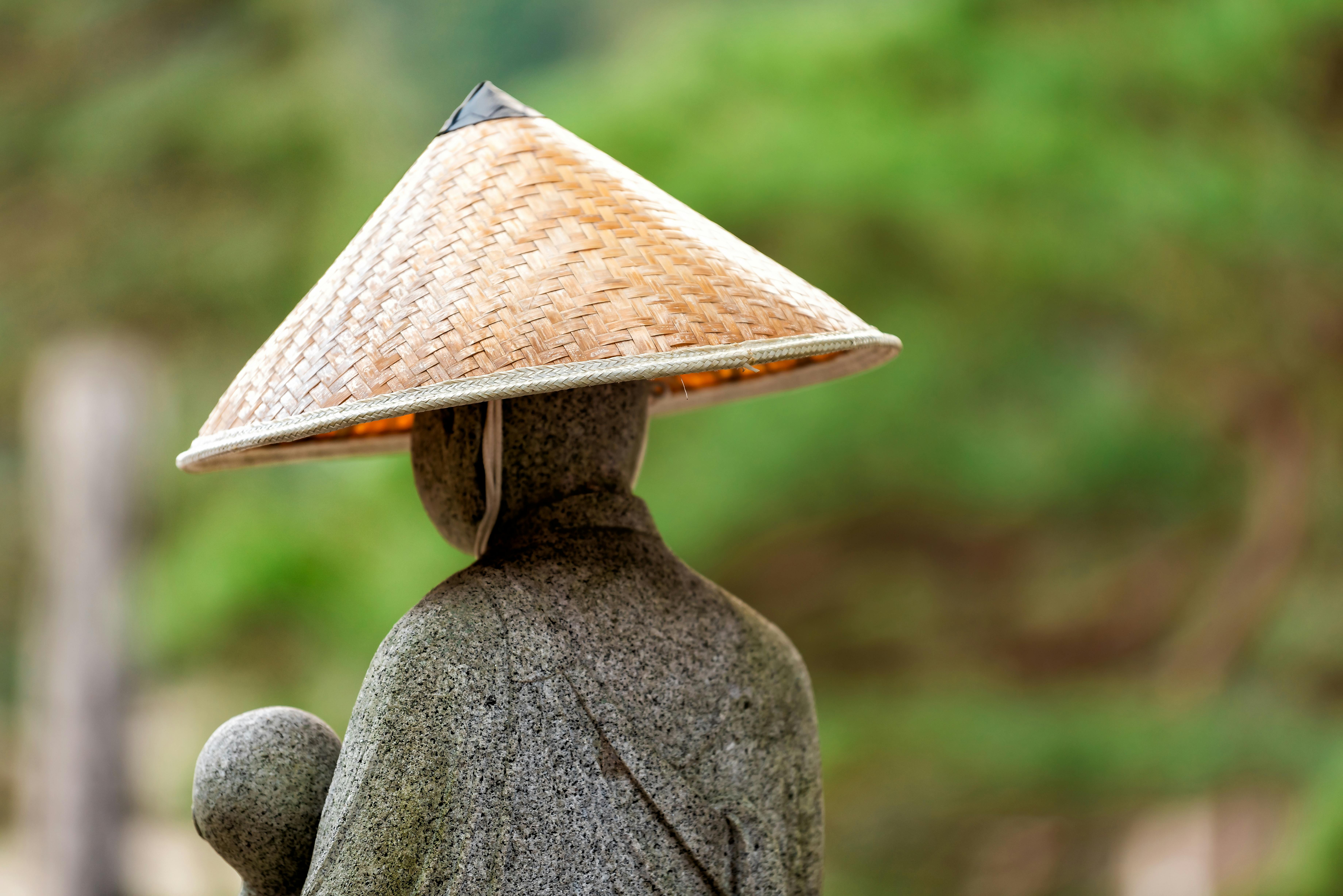 A stone statue wearing a traditional woven straw hat stands outdoors, with a blurred green natural background. The statue is seen from behind, creating a calm and serene atmosphere.