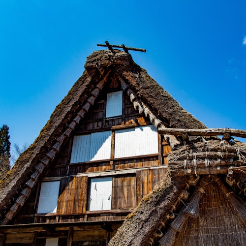 Hida Folk Village A traditional Japanese thatched-roof house with steep, triangular gables stands against a clear blue sky, surrounded by trees. The wood and straw structure shows white window coverings and rustic details.