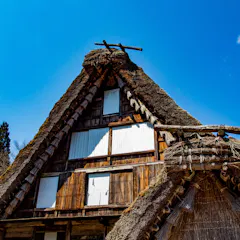 Hida Folk Village A traditional Japanese thatched-roof house with steep, triangular gables stands against a clear blue sky, surrounded by trees. The wood and straw structure shows white window coverings and rustic details.