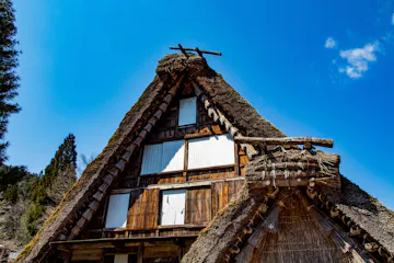Hida Folk Village A traditional Japanese thatched-roof house with steep, triangular gables stands against a clear blue sky, surrounded by trees. The wood and straw structure shows white window coverings and rustic details.