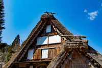 A traditional Japanese thatched-roof house with steep, triangular gables stands against a clear blue sky, surrounded by trees. The wood and straw structure shows white window coverings and rustic details.