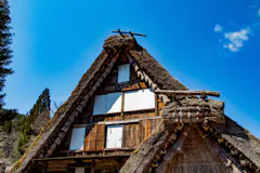 A traditional Japanese thatched-roof house with steep, triangular gables stands against a clear blue sky, surrounded by trees. The wood and straw structure shows white window coverings and rustic details.