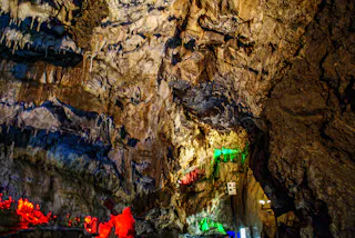A rocky cave interior with jagged walls and stalactites, illuminated by colorful lights in red, green, and blue, creating dramatic shadows and highlighting the cave’s rough texture.