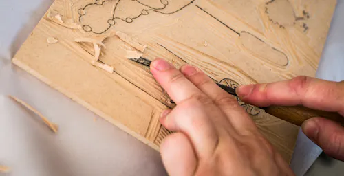 Close-up of hands carving a design into a wooden block with a chisel, with wood shavings scattered around.