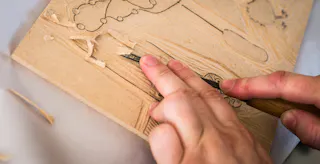 Close-up of hands carving a design into a wooden block with a chisel, with wood shavings scattered around.