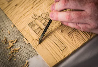 A close-up of a hand using a carving tool to cut a design of a gift box and tag into a piece of linoleum for printmaking, with wood shavings scattered around the work surface.