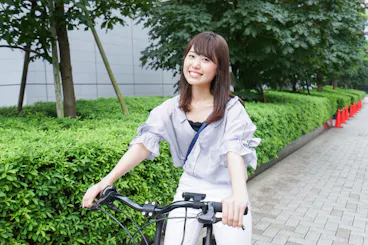 A young woman smiles while riding a bicycle on a paved path lined with green bushes and trees, with red traffic cones visible in the background.