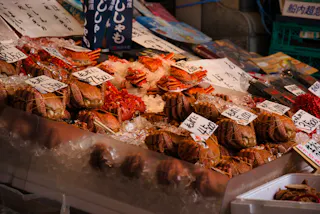 Various types of crabs are displayed on ice at a seafood market stall in Japan. Handwritten price tags in Japanese yen are visible, along with signs in Japanese above the seafood.