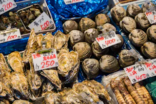A close-up of various fresh shellfish, including oysters and clams, displayed on trays at a market. Handwritten price tags in Japanese yen are visible among the seafood.