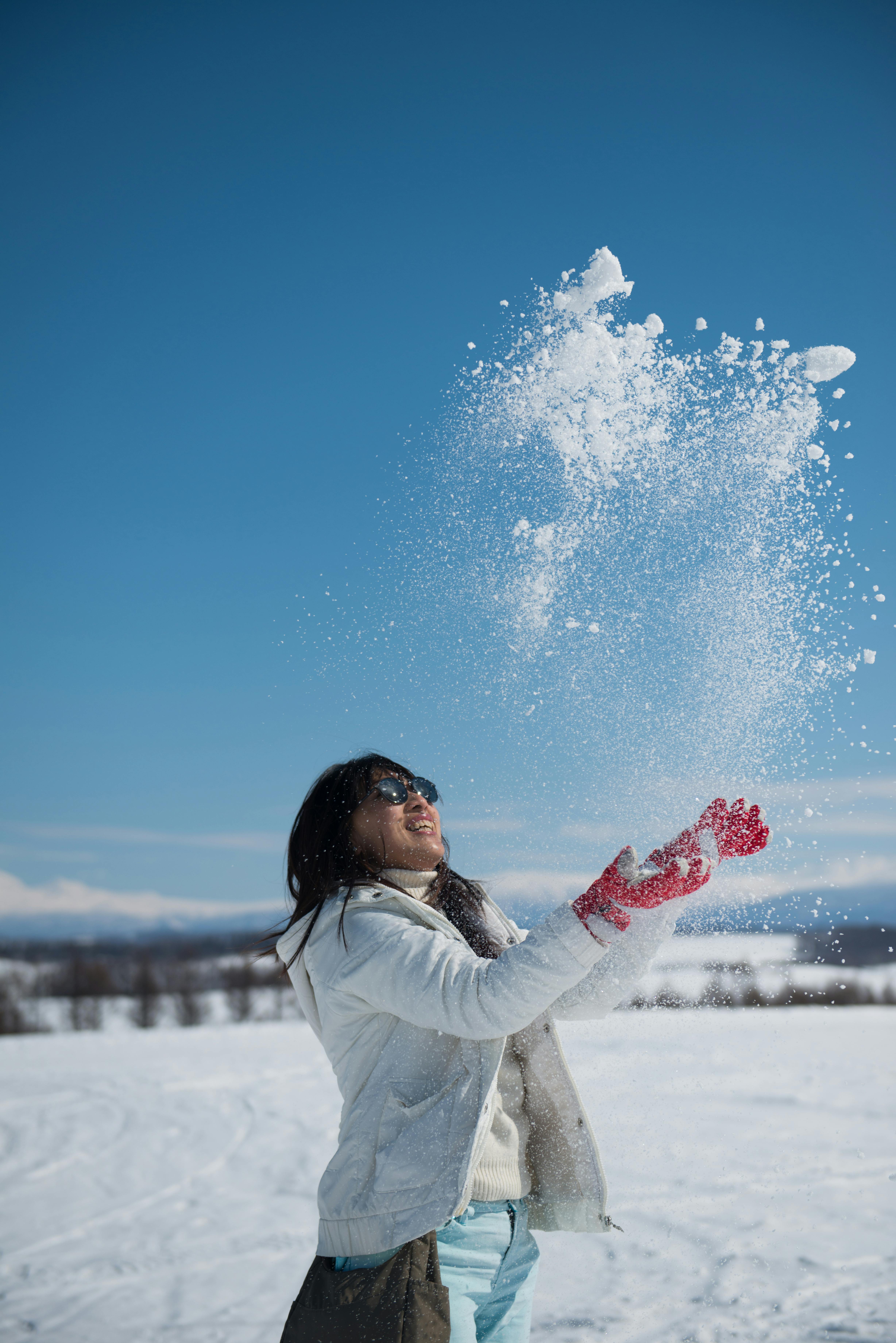 A person wearing a white jacket and red gloves tosses snow into the air and smiles under a clear blue sky in a snowy landscape.