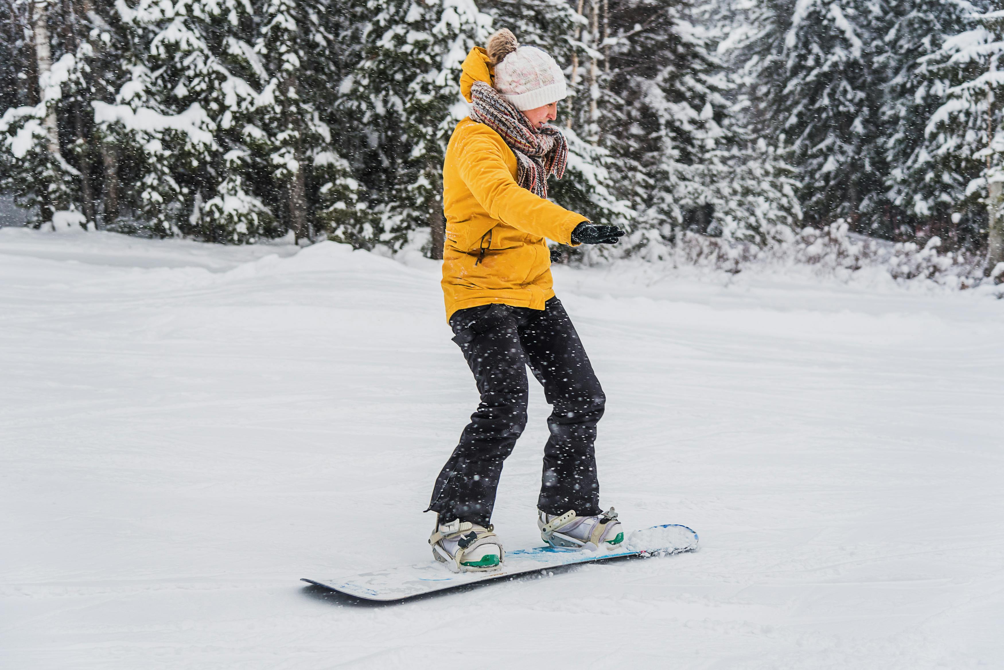 A person wearing a yellow jacket, scarf, and knit hat snowboards on a snowy slope with snow-covered trees in the background.
