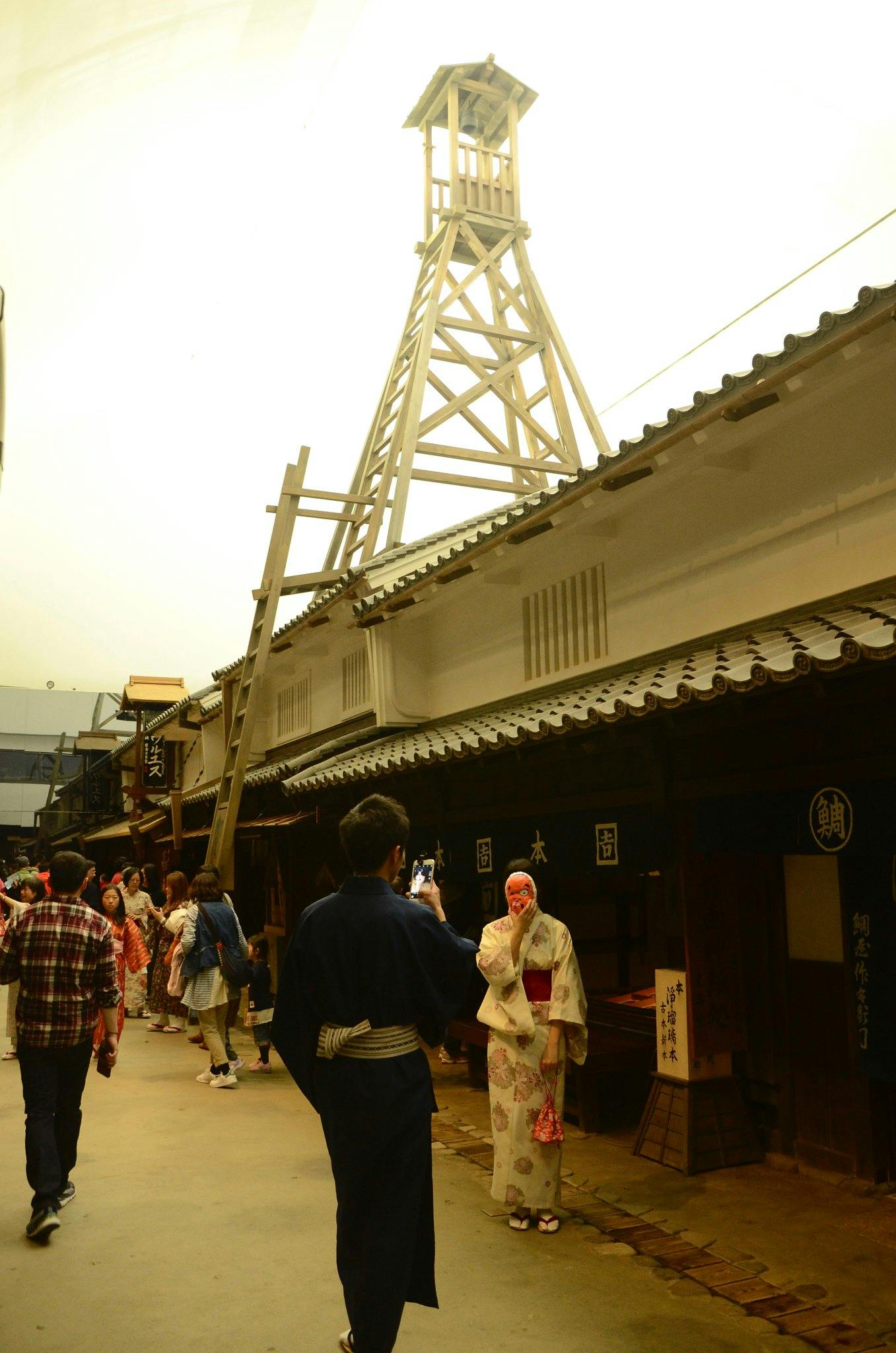 People in traditional Japanese clothing walk and pose for photos on a historical street with old wooden buildings and a tall wooden watchtower in the background, under soft indoor lighting.
