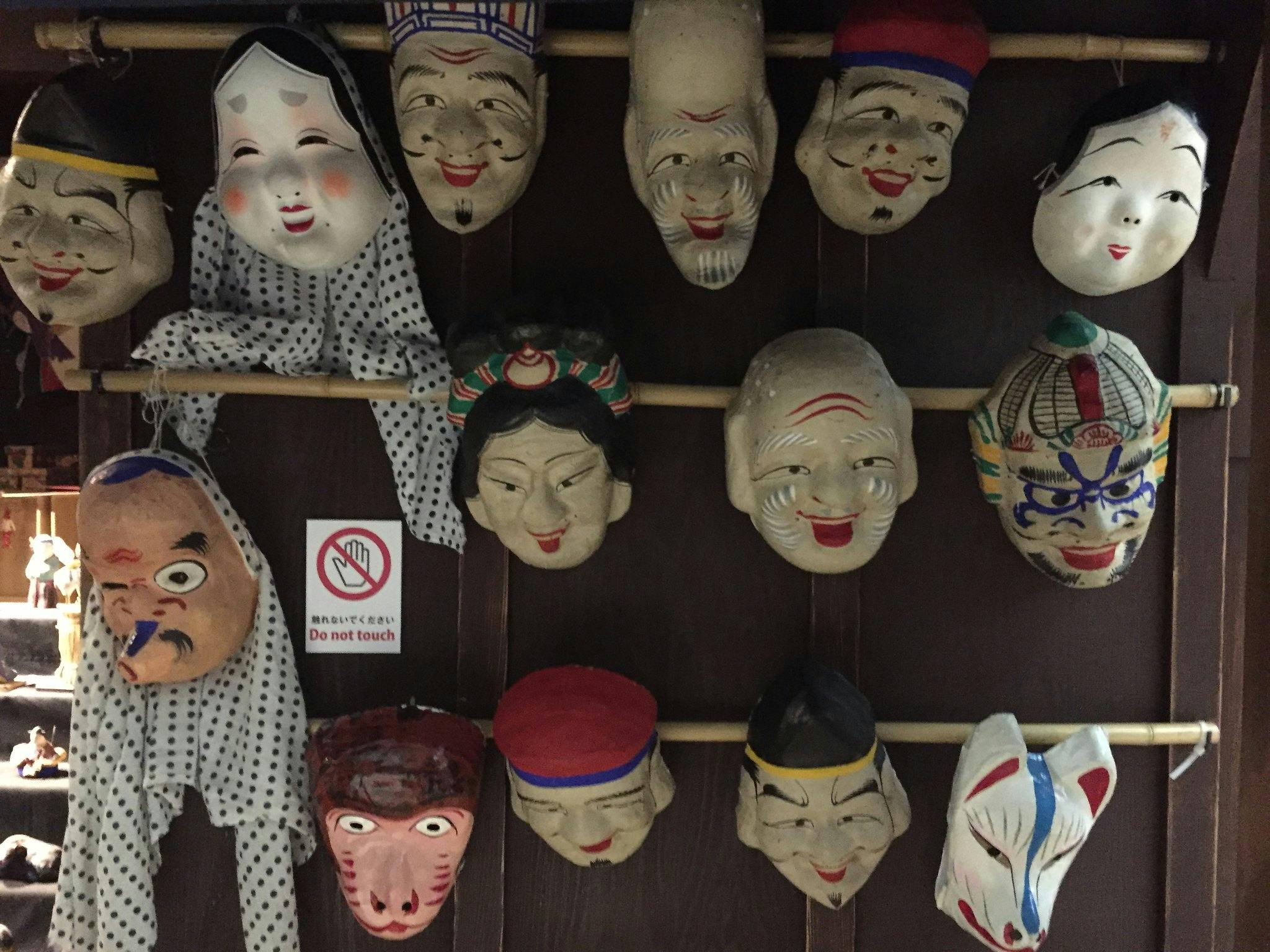 A collection of traditional Asian masks and a puppet are displayed on a wooden wall, some smiling and others with expressive faces. A “Do not touch” sign is visible among the masks.