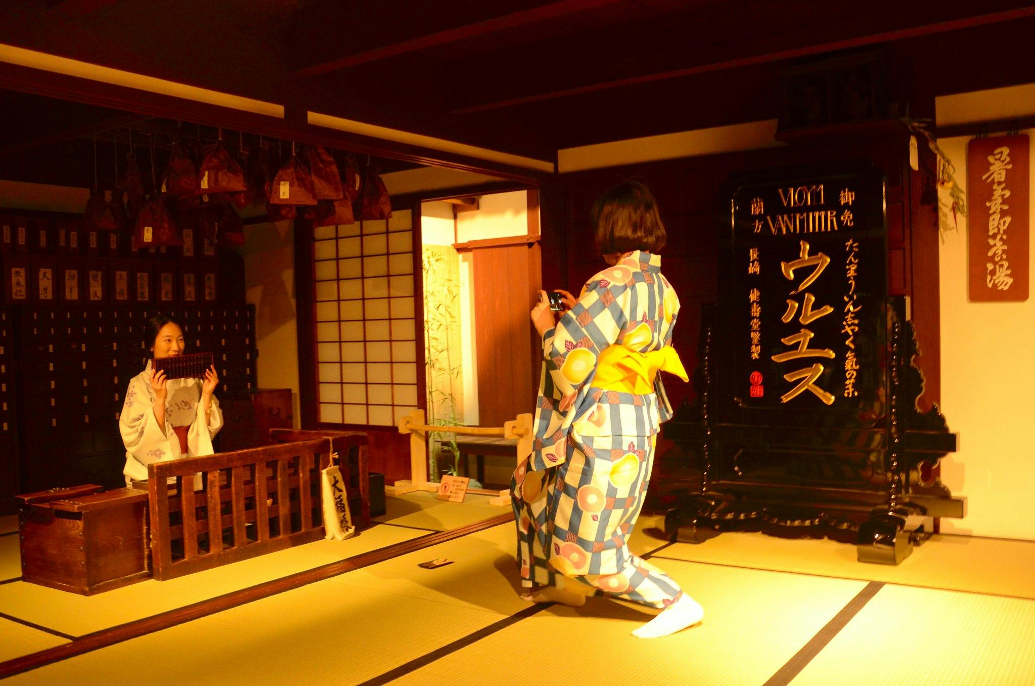 Two women in colorful kimonos are inside a traditional Japanese room. One stands taking a photo, while the other sits behind a wooden counter. The room is warmly lit and decorated with Japanese calligraphy and wooden furniture.