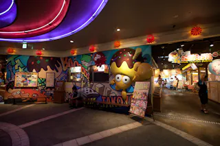 A brightly lit indoor space with colorful cartoon murals, including a large yellow character with takoyaki, and signs for a takoyaki-themed museum or attraction. Two people are visible, and menus are displayed outside.