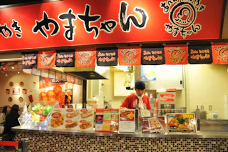 A brightly lit Japanese food stall with red signage and menus displaying various fried dishes. A staff member in a red shirt and black cap stands behind the counter. Japanese text and images decorate the stall.