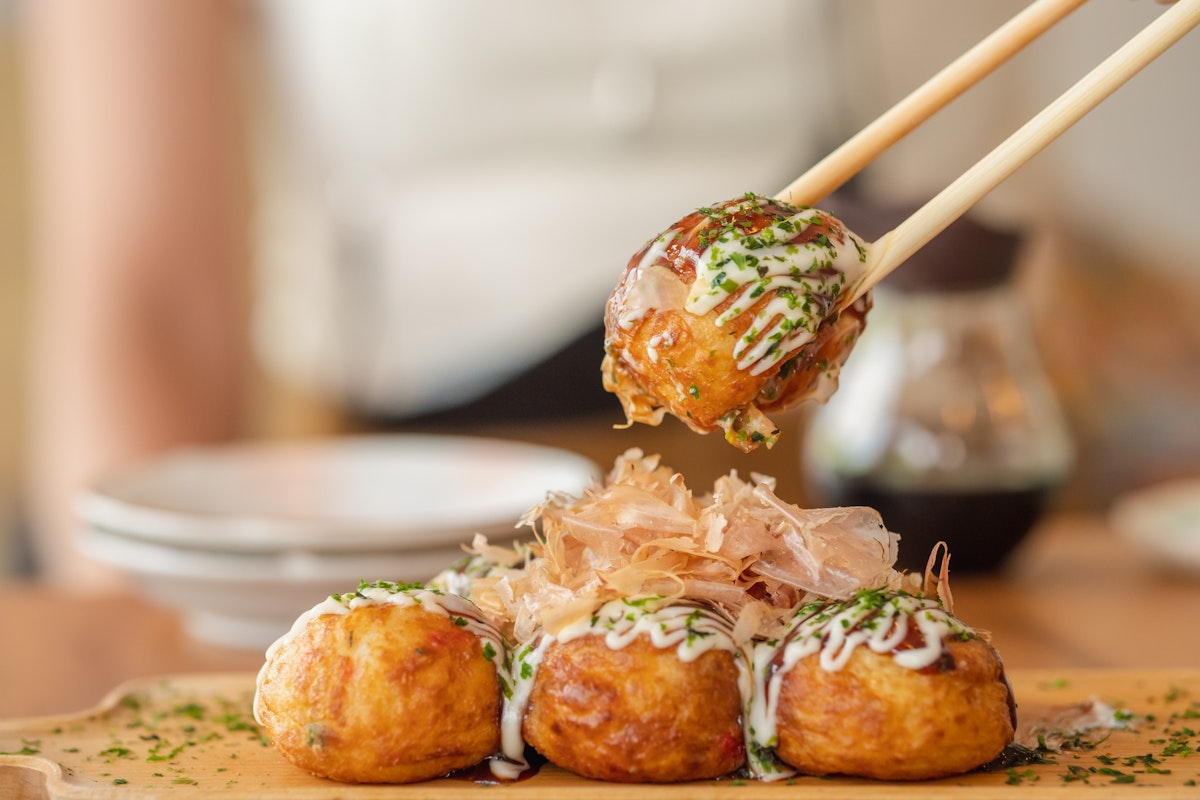Takoyaki Chopsticks holding a piece of takoyaki, a popular Japanese street food. The ball-shaped snack is topped with mayonnaise, green onions, and bonito flakes. Three more takoyaki are on a wooden plate in the background.