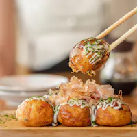 Takoyaki Chopsticks holding a piece of takoyaki, a popular Japanese street food. The ball-shaped snack is topped with mayonnaise, green onions, and bonito flakes. Three more takoyaki are on a wooden plate in the background.