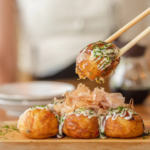 Takoyaki Chopsticks holding a piece of takoyaki, a popular Japanese street food. The ball-shaped snack is topped with mayonnaise, green onions, and bonito flakes. Three more takoyaki are on a wooden plate in the background.