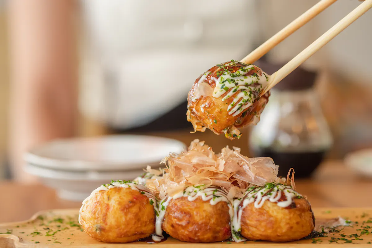 Takoyaki Chopsticks holding a piece of takoyaki, a popular Japanese street food. The ball-shaped snack is topped with mayonnaise, green onions, and bonito flakes. Three more takoyaki are on a wooden plate in the background.