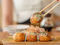 Chopsticks holding a piece of takoyaki, a popular Japanese street food. The ball-shaped snack is topped with mayonnaise, green onions, and bonito flakes. Three more takoyaki are on a wooden plate in the background.