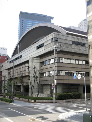 A large modern building with a curved roof and multiple stories, featuring horizontal lines and glass windows, located on a city street corner with crosswalks and trees.