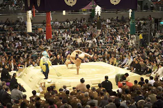 Two sumo wrestlers compete in a packed arena, surrounded by a large audience. An official and attendants are present near the circular ring, with ceremonial decorations hanging above.