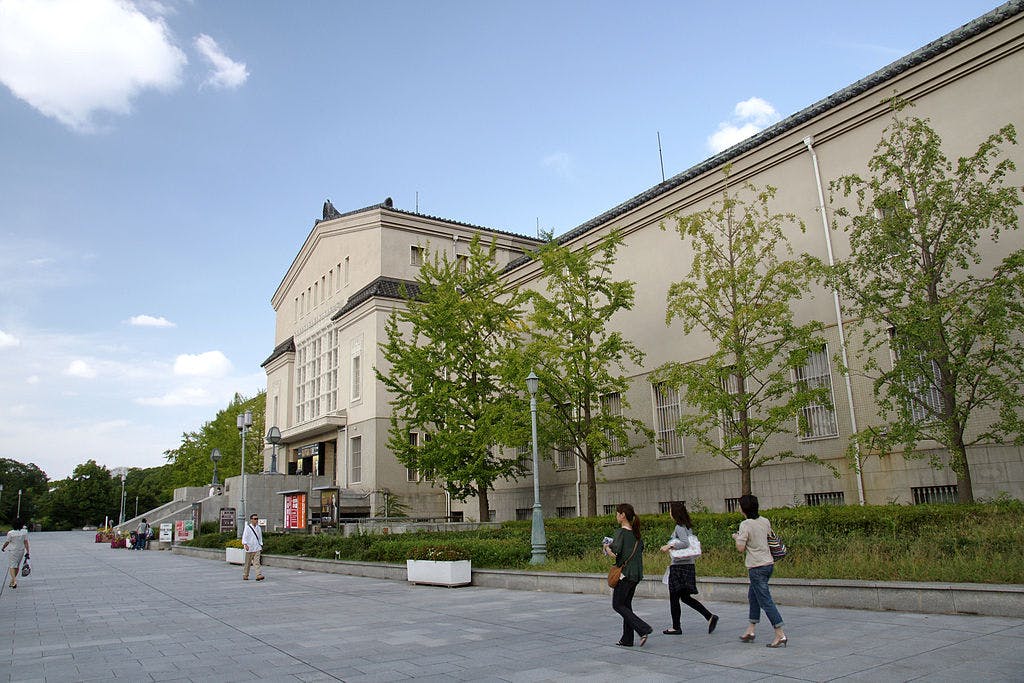 A large beige museum building with tall windows and a sloped roof stands under a blue sky. Several people walk along a wide paved path lined with green trees in front of the building.