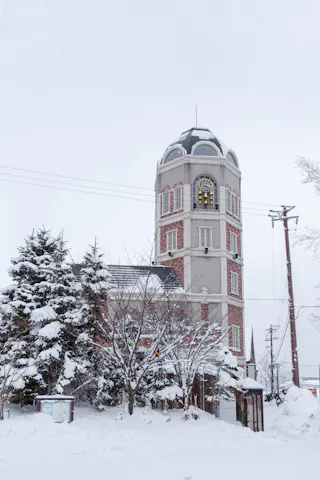 A tall, brick clock tower stands among snow-covered trees and buildings on a winter day, with thick snow blanketing the ground and rooftops. The sky is overcast, creating a quiet, serene atmosphere.