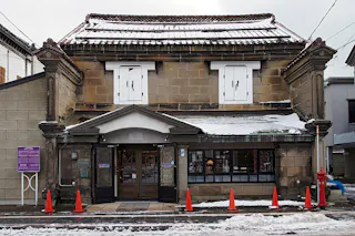 A traditional Japanese stone building with a snow-covered roof and white shutters, flanked by six orange traffic cones on a snowy street. Small icicles hang from the roof. Signs and windows are visible.