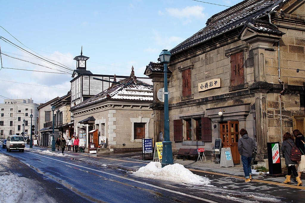A snowy street in a Japanese town with historic stone buildings, people walking on the sidewalk, and signs outside shops. Snow is piled along the roadside and rooftops.
