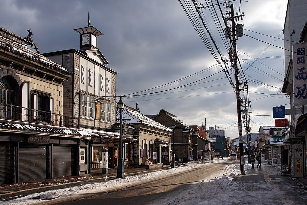 A quiet street in a Japanese town on a cloudy winter day, with snow on the ground, traditional buildings lining the road, a clock tower, and power lines stretching overhead.