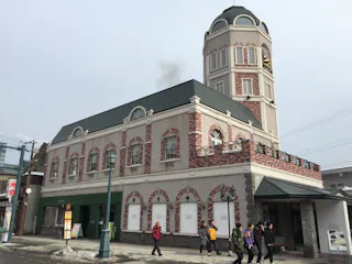 A group of people walk past a large, two-story building with a rounded clock tower and red-brick accents, on a cloudy day. The building has many windows and arched architectural details.
