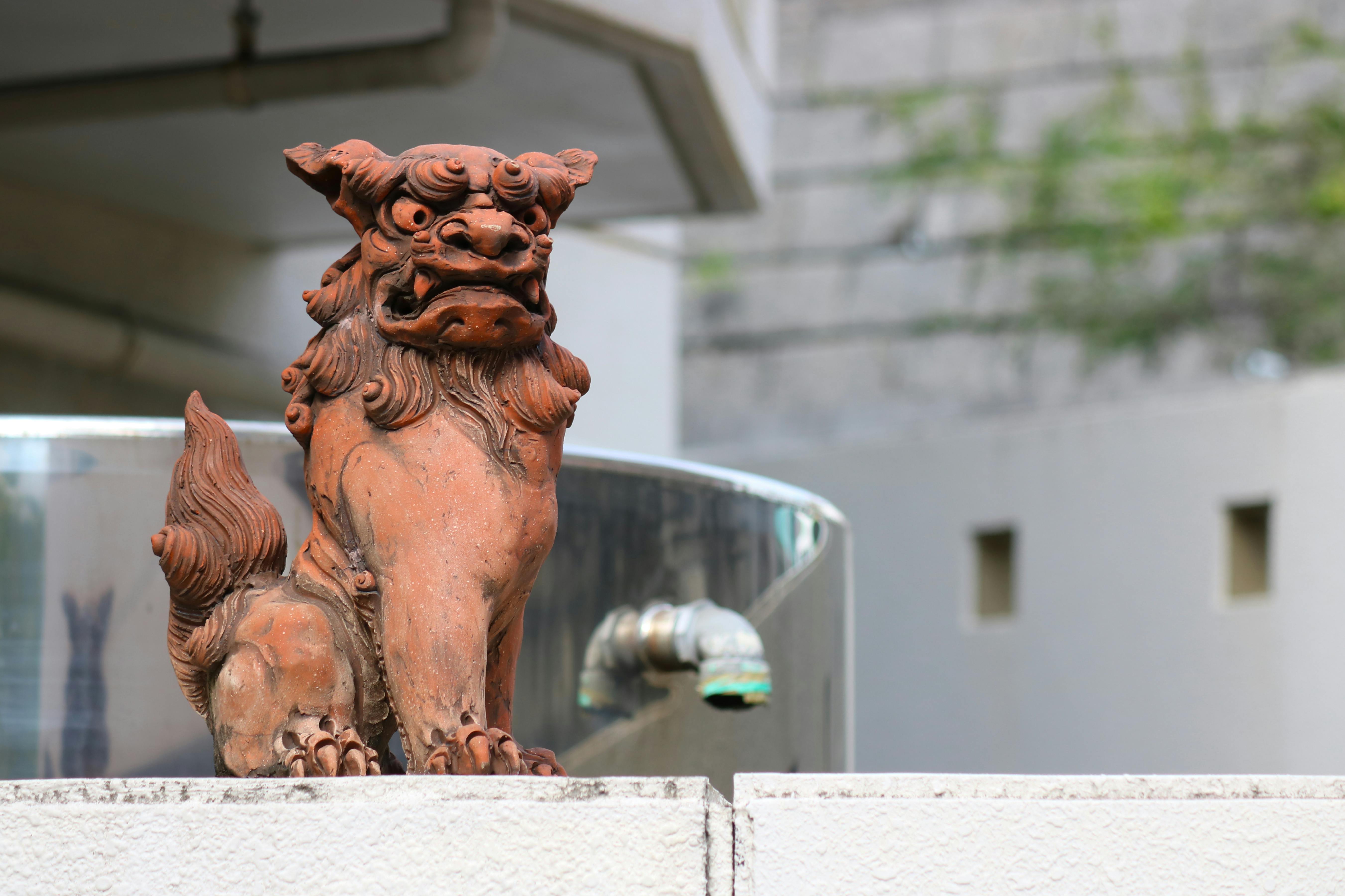 A brown Shisa lion-dog statue sits on a concrete ledge outdoors, with a curved glass railing and a water faucet in the background. The statue has a fierce expression and curly details.