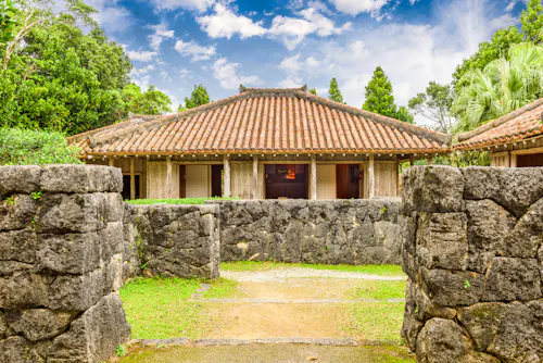 Traditional Japanese house with a tiled roof, wooden pillars, and open walls, surrounded by lush greenery and a stone wall under a partly cloudy blue sky.