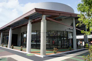 A modern, circular shopping mall building with large glass windows, white pillars, and a metal roof. There are mannequins and clothes inside, and the area has landscaped plants and patterned walkways.