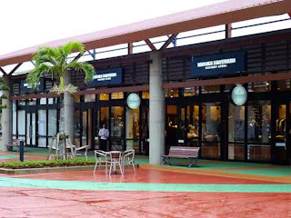 A row of factory outlet stores with large glass windows, including Brooks Brothers, under a covered walkway with a palm tree, benches, and a wet red walkway in front. A few tables and chairs are outside.