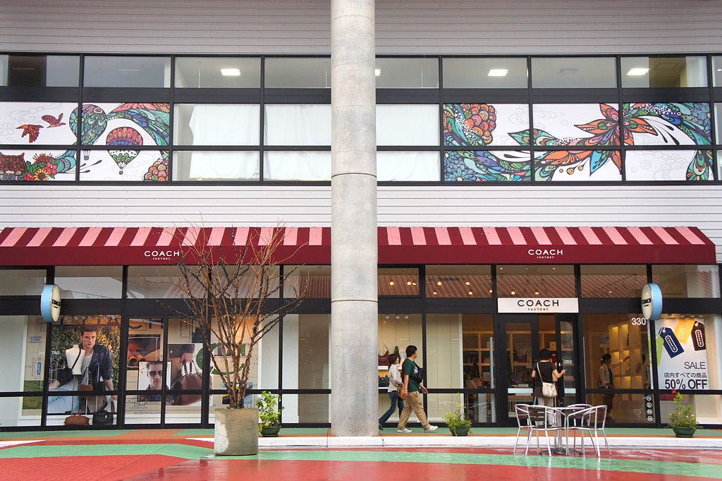 A Coach store with large glass windows, red and white striped awnings, and colorful decorative artwork above the storefront. Several people are walking by, and a small table with chairs sits outside.