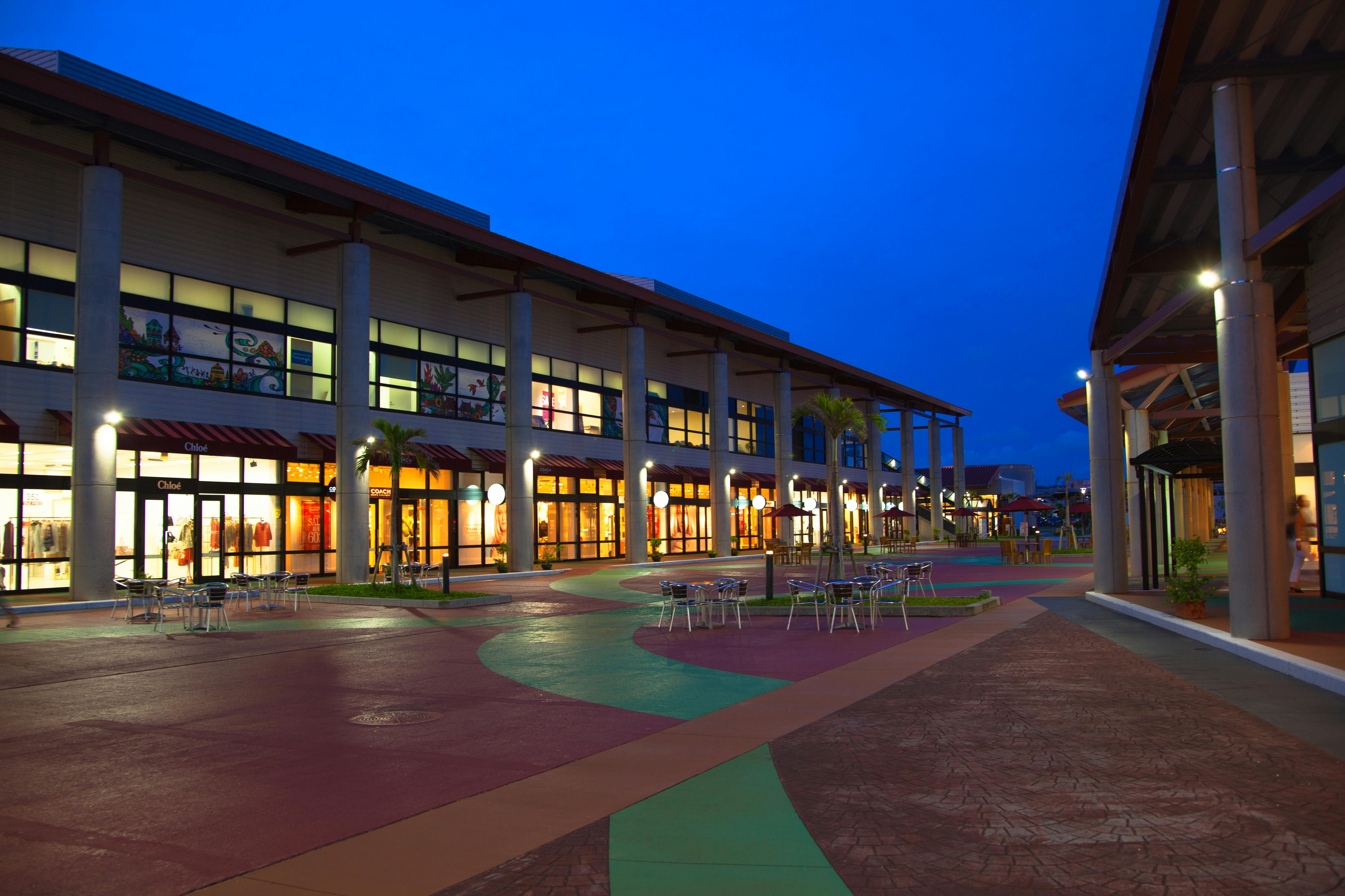 An outdoor shopping mall at dusk with two-story buildings, large windows, illuminated storefronts, colorful pathways, and empty tables and chairs arranged in the open plaza. The sky is a deep blue.