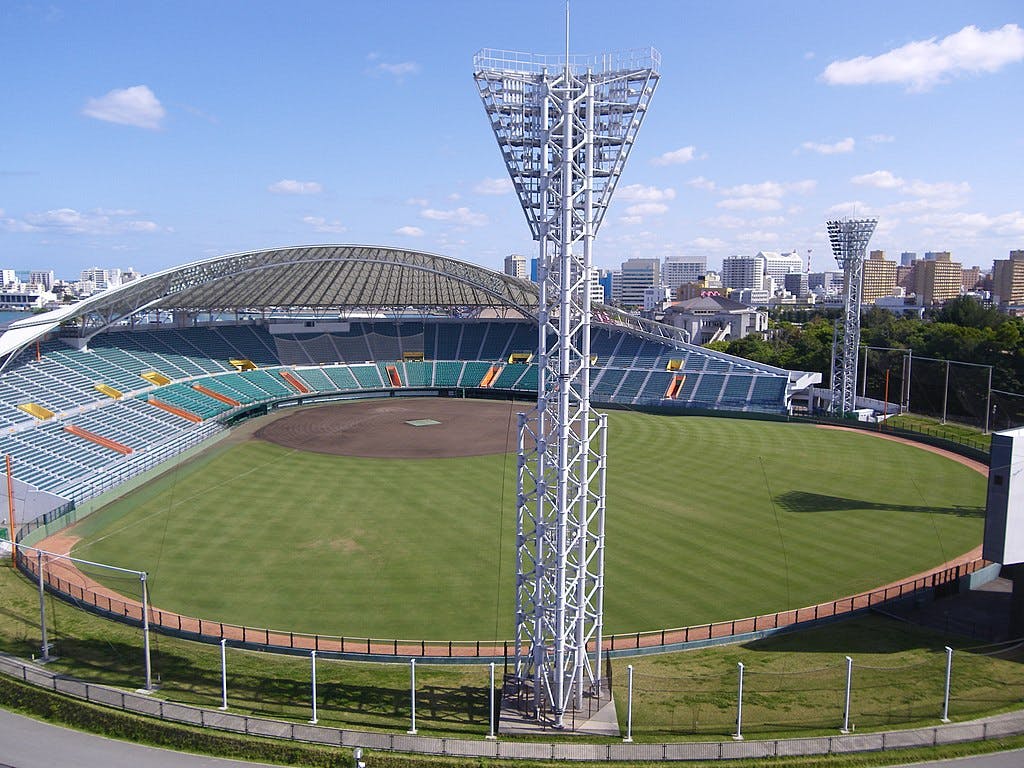 An empty baseball stadium with green seats, a grass field, and a dirt infield, surrounded by tall light towers. The stadium has a partially covered grandstand and is set against a cityscape under a blue sky.