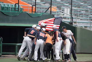 A baseball team huddles together on the field, with some wearing navy uniforms and one wearing white. An American flag is held behind them, and empty stadium seats are visible in the background.