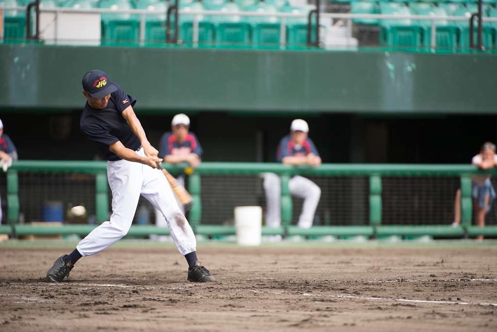A baseball player in a navy shirt and white pants swings a bat during a game, with teammates and green stadium seats visible in the background.