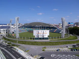 A large, modern baseball stadium with a domed roof, green field, and empty stands. Tall light towers surround the field, and there are adjacent parking lots and city buildings in the background under a clear blue sky.