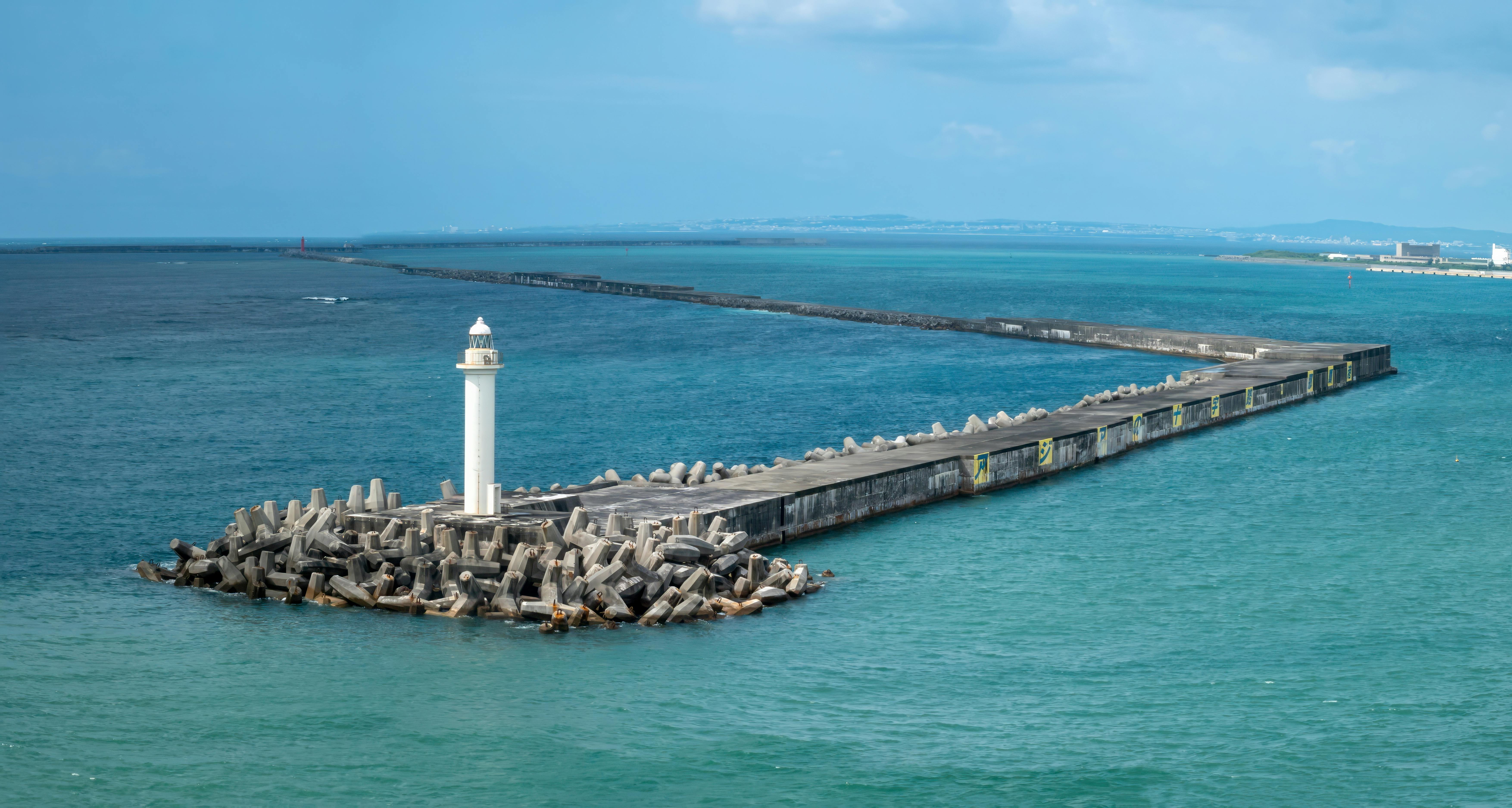 A white lighthouse stands at the end of a long, concrete pier surrounded by large wave breakers, extending into calm blue-green sea under a partly cloudy sky.