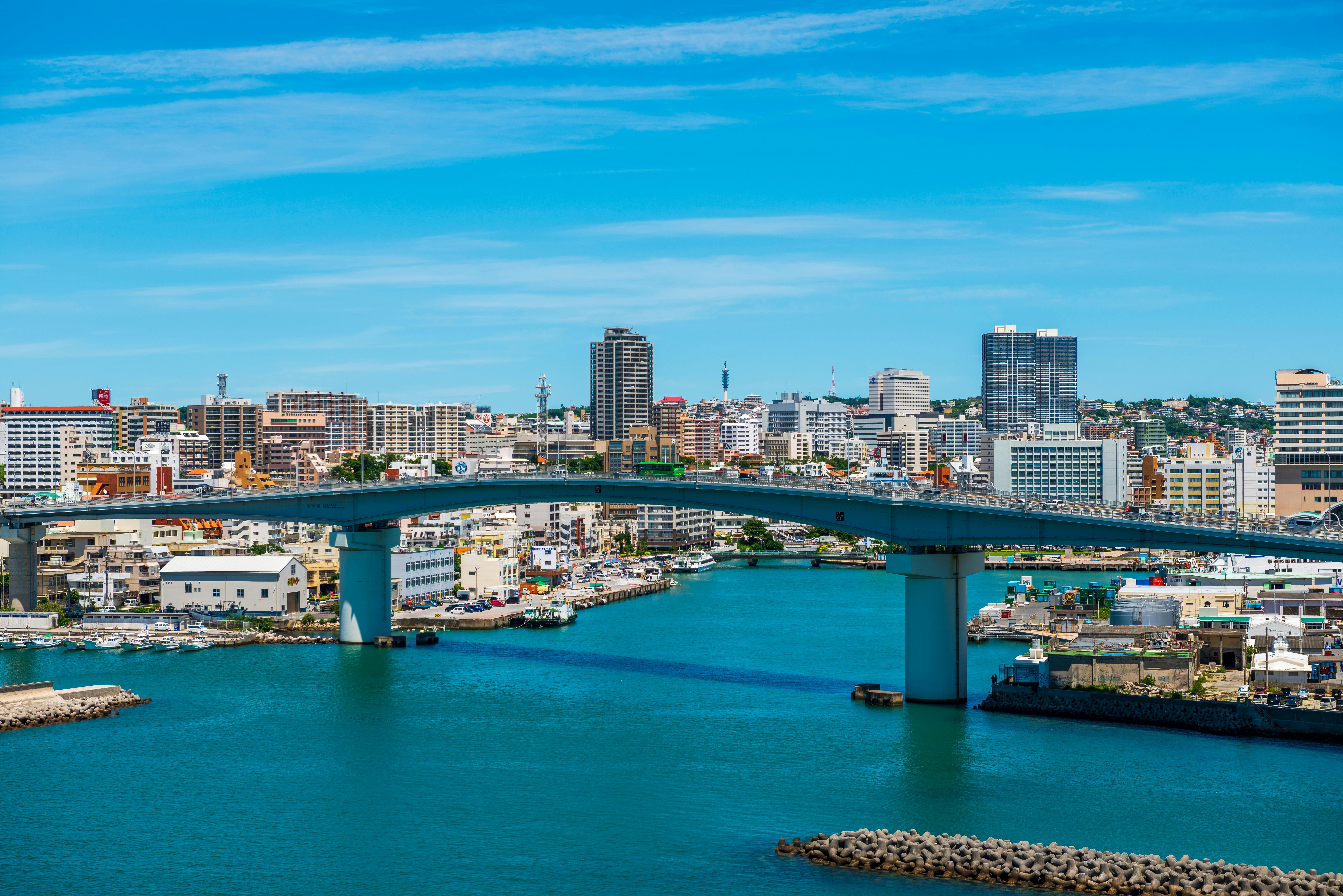 A cityscape featuring a curved bridge over turquoise water, with modern buildings and high-rises in the background under a bright blue sky.