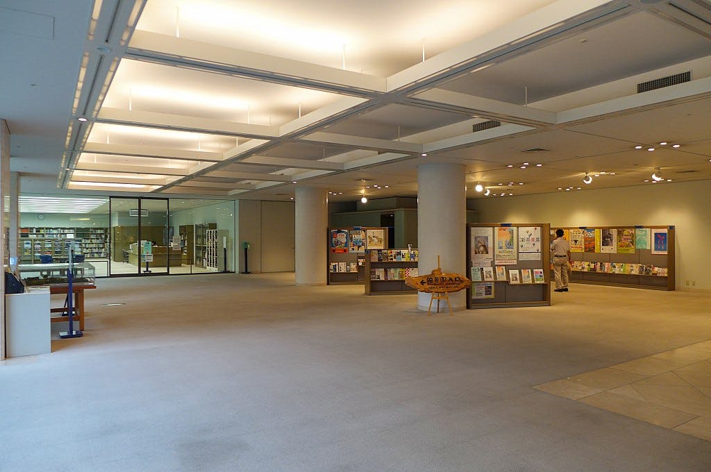 Spacious, well-lit library interior with display stands featuring books and posters. A person browses the exhibits near the wall. Glass doors lead to additional shelves and library areas in the background.