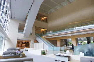 A spacious, modern building lobby with high ceilings, large windows, a central escalator, grey seating, and a few people. The interior features light-colored walls and open walkways on upper levels.