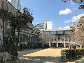 A sunny courtyard with trees and bushes, surrounded by modern buildings. Shadows from the trees fall on the sandy ground, and a few benches are visible. Blue sky with a few clouds overhead.