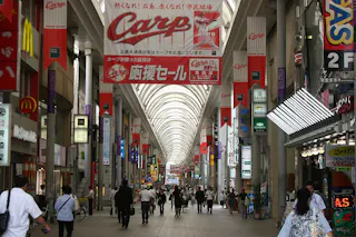 A busy covered shopping street in Japan with many people walking, surrounded by shops and tall columns. Red banners promoting the Hiroshima Carp baseball team hang from above.
