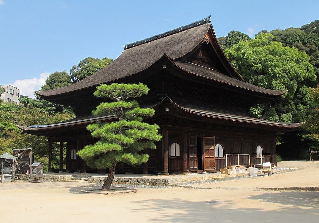 A traditional Japanese wooden temple with a curved roof stands surrounded by trees and a small pine tree in the foreground, under a bright blue sky.